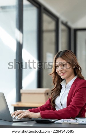Beautiful Asian businesswoman working on laptop and taking financial details from statistical graphs, charts. Successful business results in modern office with joy.