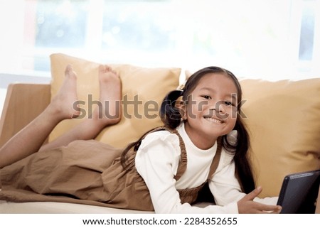 Asian girl using digital tablet while lying down on yellow sofa in living room at home, happy little kid using hi-tech devices to reading book online, child enjoy resting and watching movie on holiday