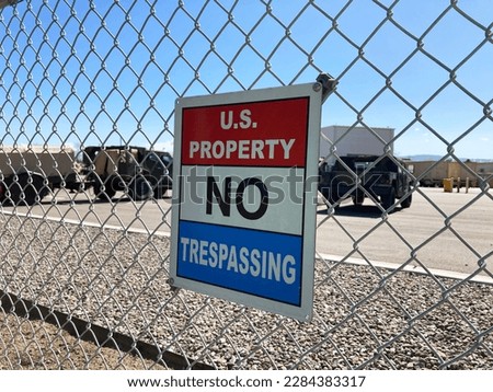 U.S. Property No Trespassing sign attached to a chain link fence. Blurred military equipment, vehicles in background.