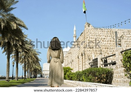 A woman in a long dress with a backpack walks along the Al Majaz embankment, Lake Khaled, Sharjah emirate. Rear view of a woman walking in the park