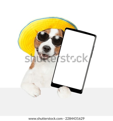Happy Jack russell terrier wearing sunglasses and summer hat holds big smartphone with white blank screen in it paw above empty white banner. isolated on white background