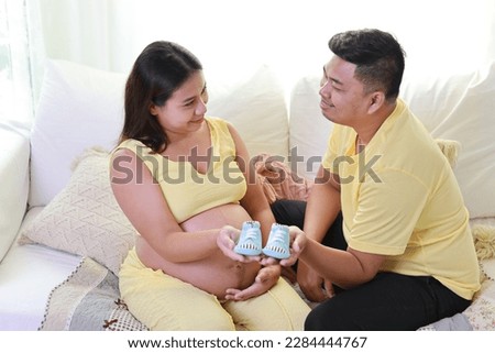 Happy asian adult couple with husband sitting and resting on sofa in living room while holding small size baby shoes and smile. Expectant mother preparing and waiting for baby birth during pregnancy.