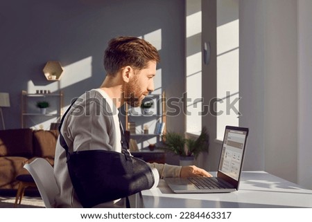 Man with broken arm working on laptop computer in office. Side view shot of businessman wearing arm splint sitting at desk in front of window working distantly at home Royalty-Free Stock Photo #2284463317