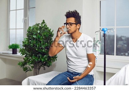 Young African American man drinking glass of water while sitting on medical bed at clinic and receiving modern intravenous anti stress vitamin therapy through sterile IV drip line infusion system Royalty-Free Stock Photo #2284463357