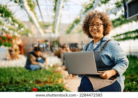 Happy female plant nursery worker using laptop and looking at camera. Copy space.