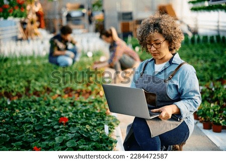 Mid adult woman working on laptop while taking care of plants in a greenhouse. 