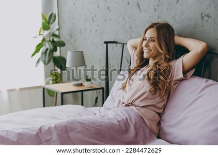 Side view young minded woman wear purple t-shirt pajama sits on bed rest relax spend time in bedroom lounge home in own room hotel wake up dream be lost in reverie good mood day. Real estate concept