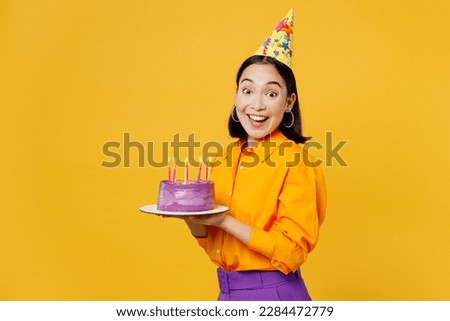 Side view happy fun smiling young woman wearing casual clothes cap hat celebrating holding purple cake with candles look camera isolated on plain yellow background. Birthday 8 14 holiday party concept