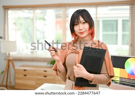 Portrait of a charming and talented young Asian female graphic designer stands in her office holding stylus and graphic tablet.