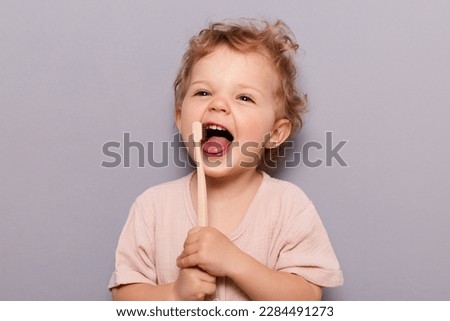 Portrait of laughing happy cheerful infant girl kid with curly blonde hair brushing her teeth, taking care of dental hygiene standing isolated over gray background. Royalty-Free Stock Photo #2284491273