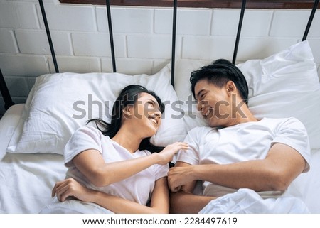 Asian new marriage couple lying down on bed and looking at each other. Attractive beautiful young man and woman in pajamas enjoy early morning activity in bedroom at home. Family relationship concept