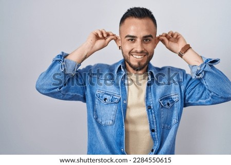 Young hispanic man standing over isolated background smiling pulling ears with fingers, funny gesture. audition problem 