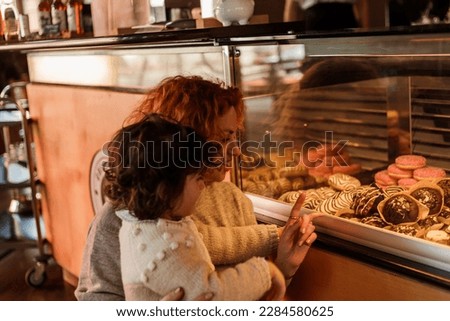 Cute little curly girl 3 years old with a young red-haired mother in the bakery at the counter chooses pastries. Cafe. Cozy.