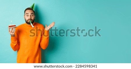 Happy birthday guy celebrating, wearing party hat, blowing wistle and holding bday cake, standing against white background.