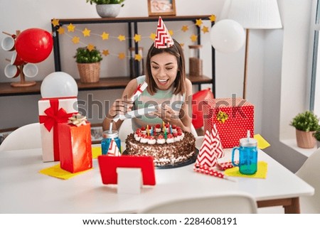Young beautiful hispanic woman celebrating online birthday opening gift at home