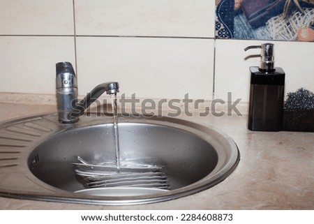Washing dishes, dishes, in the washbasin under running water, against the background of light tiles, in the kitchen. An image about housework, apartment care and cleanliness.