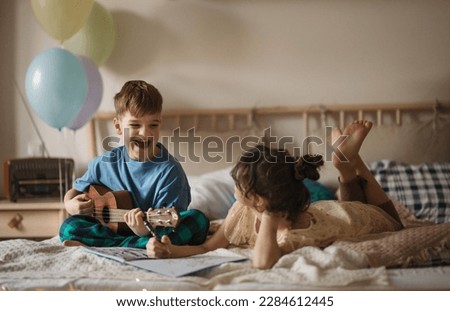 Little boy plaing on the guitar, while his sister drawing a picture.