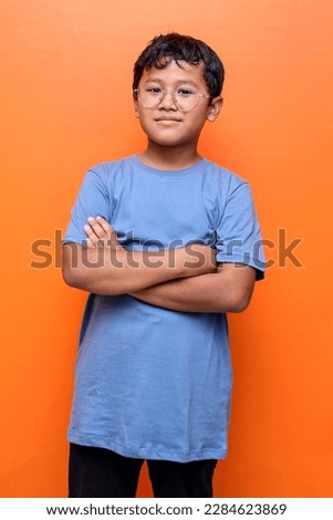 Portrait of smiling little asian boy wearing eyeglasses standing with folded arms and looking at camera, confident nerdy korean male child posing over blue studio background, copy space