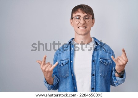 Caucasian blond man standing wearing glasses shouting with crazy expression doing rock symbol with hands up. music star. heavy music concept. 