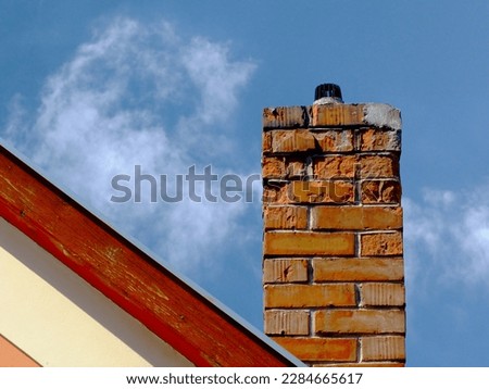 Isolated damaged clay brick chimney with weathered and spalling surface. wood trim on house gable end wall. metal flashing. white stucco exterior elevation. strong shadow. blue sky. fluffy white cloud Royalty-Free Stock Photo #2284665617