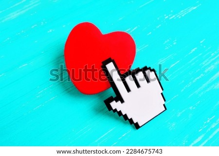 Large pixelated hand-shaped cursor touches a red heart placed on a light blue wooden background. Creative concept of connecting with others through technology or social media.