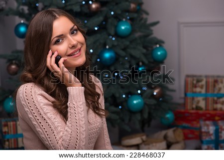 Beautiful attractive brown haired girl sitting fir tree and heap of  presents  in different  gift wrapping on white pile carpet speaking  on  phone looking at camera 