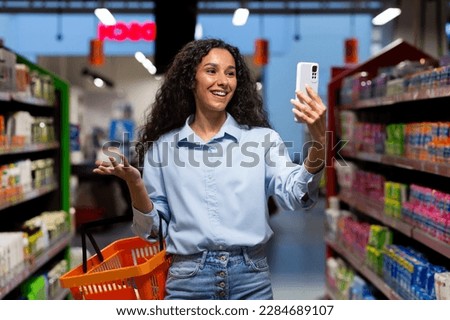 A young Latin American woman, blogger stands in a supermarket with a basket among the shelves with goods and talks on a video call from the phone, consults, asks what to buy, smiles. Royalty-Free Stock Photo #2284689107