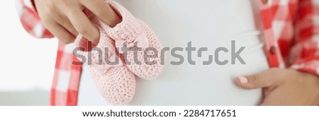 Close-up of pregnant woman holding pink baby booties on belly. Happy and healthy pregnancy, maternity, preparation and expectation concept
