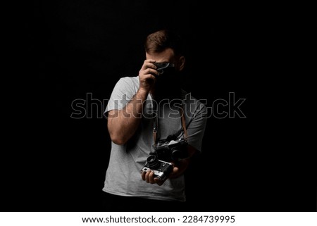 Bearded professional photographer in a grey t-shirt with a bunch of different cameras in a hands and on a shoulder looking on a camera and ready for make a good shoot.
