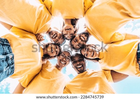 Multi racial group of young people standing in circle and smiling at camera with yellow uniform - Volunteers take picture - Happy diverse friends having fun hugging together outdoor - Low angle view