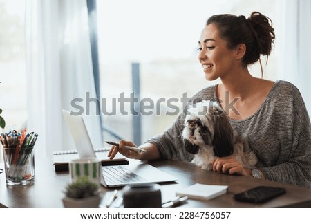 Attractive young female freelancer working on laptop from her home and having her pet dog in her lap to keep her company. Royalty-Free Stock Photo #2284756057