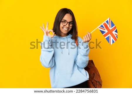 Young latin woman holding an United Kingdom flag isolated on yellow background showing ok sign with fingers