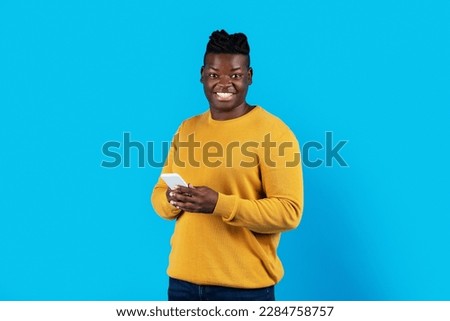 Portrait Of Millennial Black Man Holding Mobile Phone And Looking At Camera, Cheerful Young African American Guy Enjoying Mobile Communication, Standing Isolated Over Blue Background, Copy Space
