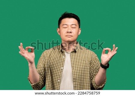 Stress relief concept. Asian man meditating with closed eyes, keeping calm on green background, studio shot. Middle aged korean male practicing yoga, searching for inner balance