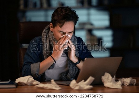 Unhealthy sick young man with scarf around his neck employee working late at office, guy sitting at workdesk, looking at laptop screen and sneezing, using napkin, got flu, coronavirus, copy space Royalty-Free Stock Photo #2284758791