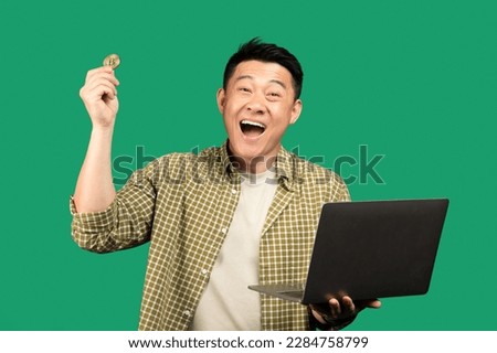 Overjoyed asian businessman holding laptop and bitcoin coin, posing over green studio background, looking at camera with excitement. Cryptocurrency, economics, saving, app for modern business