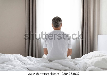 Depression, midlife crisis, mental breakdown. Back view of grey-haired middle aged man wearing pajamas sitting alone on bed after waking up at home, looking through window, copy space Royalty-Free Stock Photo #2284758839