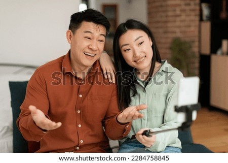 Smiling korean married spouses with happy faces looking at cellphone webcamera, bloggers recording videoblog on phone at home. Video live streaming Royalty-Free Stock Photo #2284758845