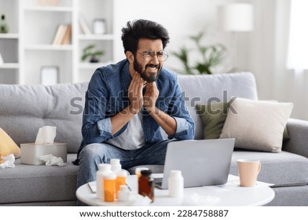 Seasonal Cold And Telemedicine. Sick young indian man suffering from sore throat while sitting on couch at home, Ill eastern man touching neck with hands and frowning, having acute pain, copy space Royalty-Free Stock Photo #2284758887