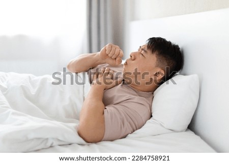 Unhealthy sick middle aged asian man wearing pajamas lying in bed and coughing, touching his chest, home interior, side view, copy space. Lung cancer, pneumonia, smokers cough Royalty-Free Stock Photo #2284758921