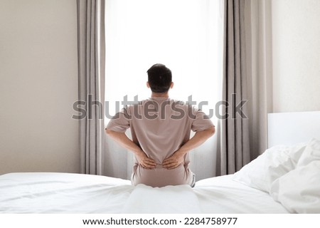 Tired man suffering from back pain after sleeping in bedroom, waking up in morning, sitting on bed and touching his lower back, experiencing muscle strain, wearing pajamas, copy space, back view Royalty-Free Stock Photo #2284758977