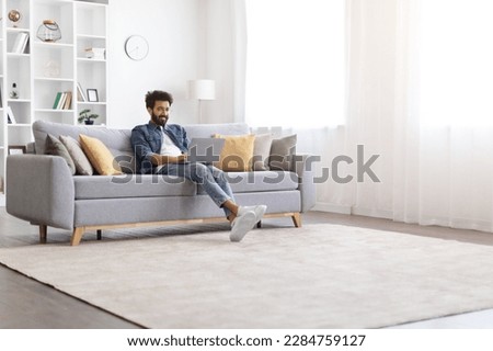Handsome indian man working on laptop while sitting on couch at home, happy young eastern male freelancer typing on computer keyboard, enjoying remote work and online communication, copy space Royalty-Free Stock Photo #2284759127