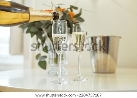 Closeup Of Sparkling Wine Bottle And Alcohol Pouring In Two Champagne Glasses Standing On Table Near Bucket And Vase With Flowers Indoors. Drinks Serving Concept. Cropped Shot, Selective Focus Royalty-Free Stock Photo #2284759187