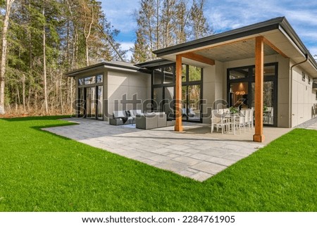 Exterior image of a contemporary home with flat roof and brown trim and lush green grass blue sky forest in the background furnished patio for outdoor dining and lounging Royalty-Free Stock Photo #2284761905