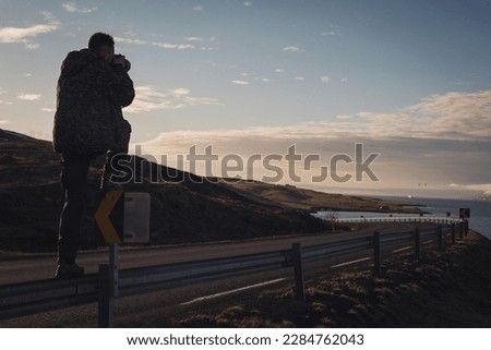 Nature photographer in the action. Man silhouette above a road, evening hilly landscape. Silhouette of Photographer on sunset background Twilight and photographer on viewpoint taking photo in Iceland.