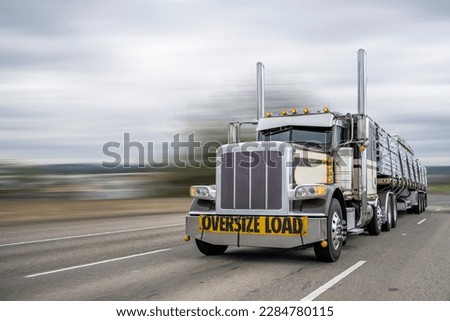 Classic beige big rig semi truck tractor with oversize load sign on the bumper and turned on light transporting transporting cargo on flat bed semi trailer driving on the wide highway road