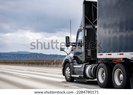 Day cab local freight black big rig industrial semi truck tractor with roof spoiler transporting commercial cargo in tented dry van semi trailer running on the highway road with clouds sky Royalty-Free Stock Photo #2284780171