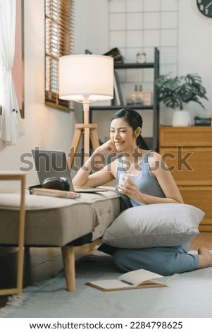 Millennial Asian girl sits on sofa in living room, studying on laptop making notes.The concentrated young woman works on computer and writes in her notebook, taking online course or training at home