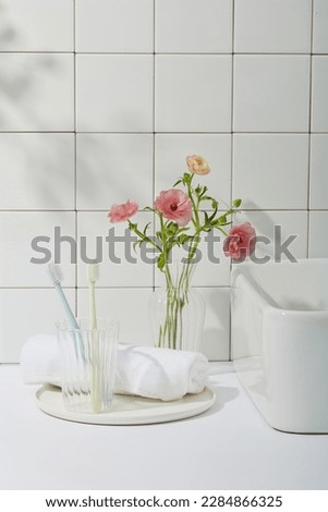 Beauty photo with bathroom concept. Toothbrushes in glass cup, white towel on round dishes, one corner of the washbasin and pink flower vase on tile wall background. Front view, copy space