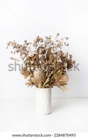 Beautiful dried flower arrangement of Australian native banksia, eucalyptus leaves, red leucadendrons and delicate white flowers, in a white vase on a table with a white background. Royalty-Free Stock Photo #2284870495